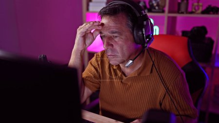 Concentrated mature man wearing headset using computer in a neon-lit gaming room at home.