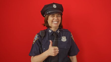 Una sonriente mujer policía hispana de mediana edad dando un pulgar en contra de un fondo rojo aislado.