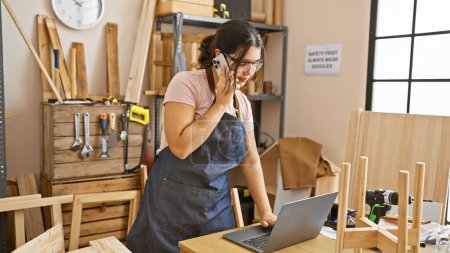Foto de Mujer hispana joven en delantal usando laptop y teléfono en un taller de carpintería - Imagen libre de derechos
