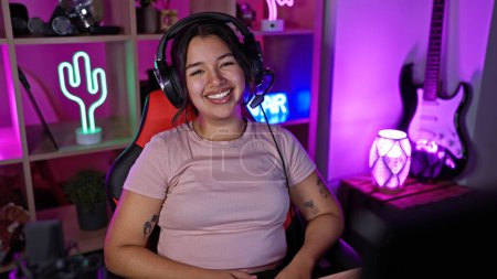 Portrait of a smiling young hispanic woman wearing headphones in a vibrant gaming room at night.