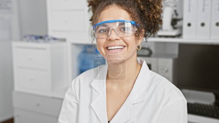 Una joven hispana sonriente con el pelo rizado usando gafas de seguridad y una bata de laboratorio en una clínica.