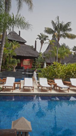 Téléchargez les photos : Une femme se promène au bord de la piscine dans un luxueux complexe bali entouré de palmiers et de chaises longues. - en image libre de droit