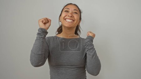 Foto de Joven mujer hispana alegre celebrando con los puños cerrados contra una pared blanca lisa en una capota gris casual. - Imagen libre de derechos