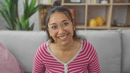 Téléchargez les photos : Une jeune femme hispanique souriante avec des lunettes assises à la maison qui dégage une ambiance décontractée et accessible. - en image libre de droit