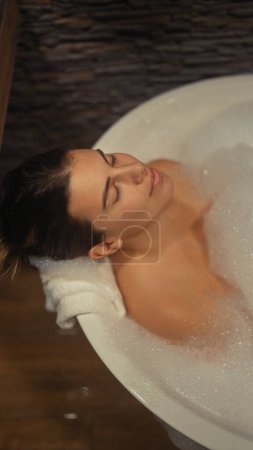 Eine ruhige junge Frau genießt ein entspannendes Schaumbad in einem gemütlichen, holzbetonten Badezimmer.