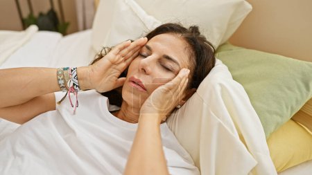 Foto de Una mujer hispana de mediana edad con el pelo rizado parece estresada o con un dolor de cabeza en un ambiente acogedor dormitorio. - Imagen libre de derechos