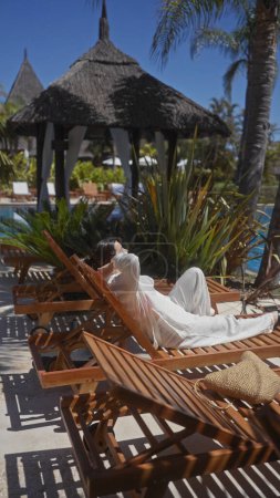 Téléchargez les photos : Une femme détendue se prélassant sur un transat en bois dans un luxueux complexe bali, entouré de végétation tropicale. - en image libre de droit