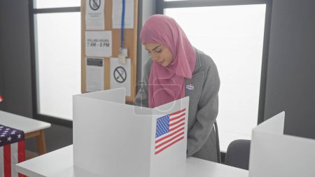Una mujer en un hiyab emitiendo su voto en un centro de votación americano con cabinas de privacidad y banderas.