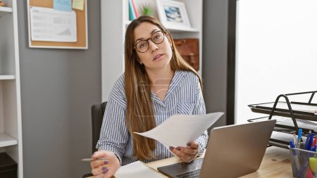 Eine professionelle junge Frau begutachtet Dokumente am Schreibtisch ihres Büros, trägt eine Sportbrille und lange brünette Haare im gestreiften Hemd..