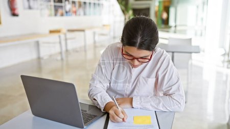 Photo for A focused young hispanic woman working and writing in a modern office setting, embodying professionalism and concentration. - Royalty Free Image
