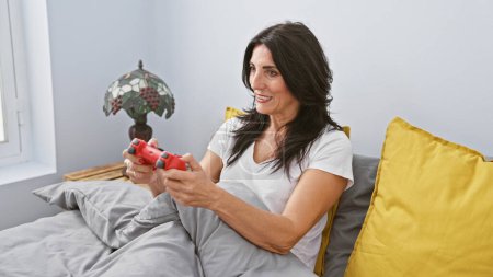 Foto de Una morena de mediana edad en un dormitorio, sonriendo mientras juega con un controlador de juego rojo - Imagen libre de derechos