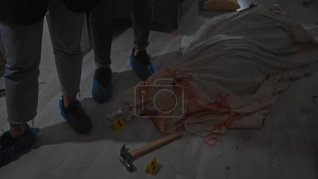 Téléchargez les photos : Deux enquêteurs en couvre-chaussures analysent une scène de crime ensanglantée avec des étiquettes numérotées et un marteau à l'intérieur. - en image libre de droit