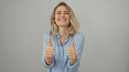 A cheerful young adult caucasian woman with blonde hair, giving thumbs up in a blue shirt isolated on white background.