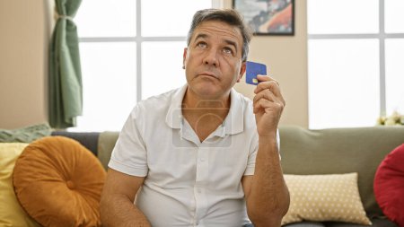 Photo for Pensive middle-aged man holding credit card sitting in living room contemplating - Royalty Free Image