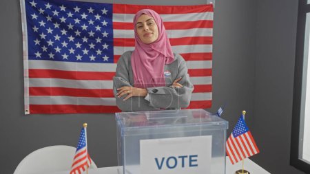 Eine attraktive junge hispanische Frau im Hidschab steht mit verschränkten Armen in einem amerikanischen Wahlzentrum mit Fahnen und einer "Wahlkabine"