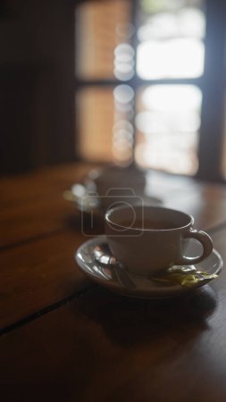 A cozy cafe scene with warm lighting featuring a white cup of coffee on a saucer with a spoon and sugar packet.