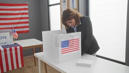 Una mujer madura con un traje vota atentamente en el centro de votación con una bandera de nosotros en el fondo