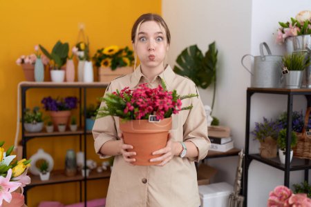 Young caucasian woman working at florist shop holding plant pot puffing cheeks with funny face. mouth inflated with air, catching air. 