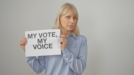 A middle-aged woman with blonde hair, standing against a white backdrop, holds a sign about voting rights.