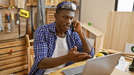 Photo for An african american man talking on the phone in a carpentry workshop with a laptop, safety glasses, and woodworking tools - Royalty Free Image