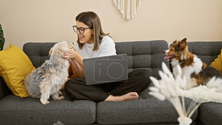 Hispanic woman working on laptop with two dogs in a cozy living room, depicting a comfortable work-from-home setting.