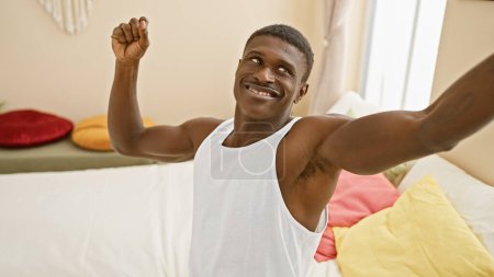 A cheerful african man in a white tank top stretching in a colorful bedroom with pillows and sunlight.