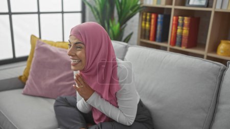 Photo for A smiling young woman with a pink hijab seated on a grey couch in a cozy living room. - Royalty Free Image