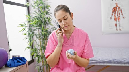 Brunette woman in pink scrubs using phone while holding massage ball in a rehabilitation clinic.