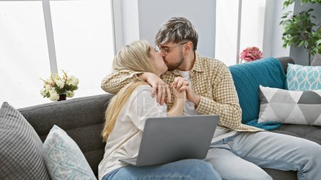 A loving couple kissing on a cozy sofa with a laptop in a modern living room