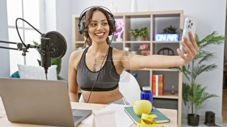 Una mujer sonriente con auriculares se toma una selfie mientras podcasting en un moderno estudio de radio interior.