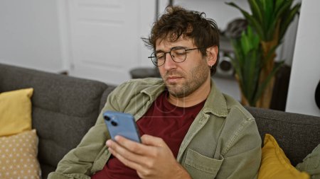Hispanischer Mann mit Bart und Brille mit Smartphone auf Couch drinnen.