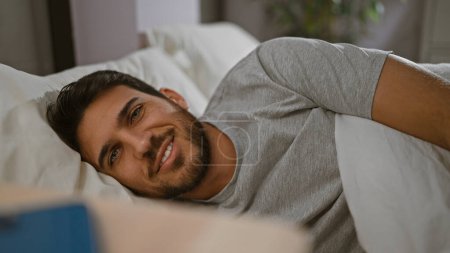 Téléchargez les photos : Un jeune homme hispanique avec une barbe sourit dans un lit, donnant un sentiment de détente et de confort dans un cadre familial. - en image libre de droit