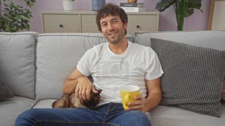 Photo for A smiling young hispanic man pets a cat while sitting on a living room couch with a cup in hand. - Royalty Free Image