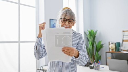 Eine glückliche Frau mittleren Alters mit Brille und grauen Haaren feiert Erfolg in einem modernen Büroumfeld.