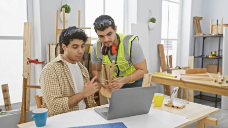 Photo for Two men discuss a woodcraft project in a well-equipped carpentry workshop, referencing a laptop. - Royalty Free Image
