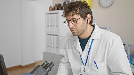 Handsome bearded man wearing glasses and a white lab coat with wireless earphones sits thoughtfully in a clinic.