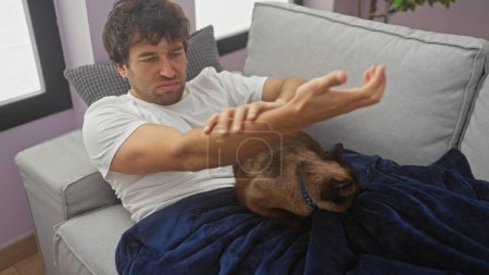 Photo for Young hispanic man indoors in a living room experiencing arm pain while sitting on a couch with his pet cat under a blanket. - Royalty Free Image