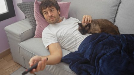 A relaxed young man lounges on a sofa, petting a siamese cat while holding a remote in a cozy living room.