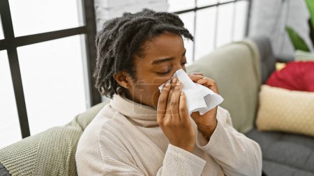 Eine junge Frau mit Dreadlocks pustet ihre Nase auf einem Sofa drinnen, was auf Krankheit oder Allergien hindeutet.