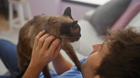 Foto de Joven hombre hispano vinculándose con su gato siamés en un acogedor hogar interior - Imagen libre de derechos