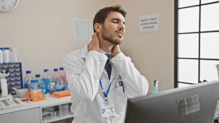 Photo for Hispanic man in lab coat feeling neck pain inside hospital laboratory with computer and medical equipment - Royalty Free Image