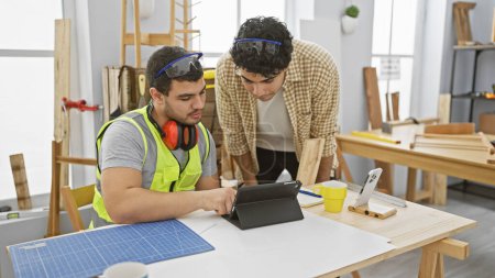 Two men reviewing plans on a tablet in a bright carpentry workshop, wearing casual and safety gear.