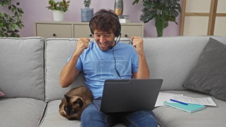 Photo for Excited man with headphones celebrates at laptop with pet cat on sofa indoors - Royalty Free Image