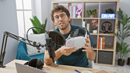 Foto de Hombre hispano sosteniendo auriculares vr en un estudio de podcast con micrófono y señal en el aire. - Imagen libre de derechos