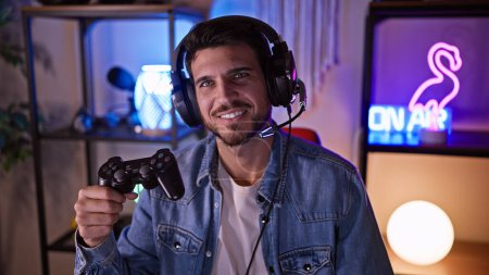 A smiling young hispanic man with a beard wearing headphones holds a game controller in a neon-lit gaming room at home.