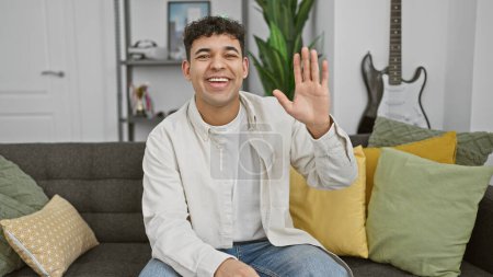 Photo for Handsome young man waving hello in a stylish living room, expressing friendliness. - Royalty Free Image