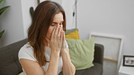 Reife Frau in Bedrängnis mit Händen, die das Gesicht verdecken, wenn sie zu Hause auf der Couch sitzt und Gefühle der Traurigkeit oder Sorge vermittelt.