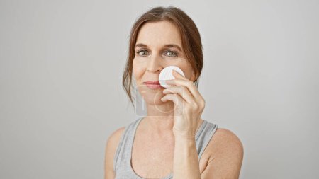 Mature woman applies skincare routine holding cotton pad on her face against white background