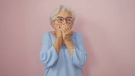 A surprised senior woman with grey hair and glasses standing against a pink background, covering her mouth with hands in a blue sweater.