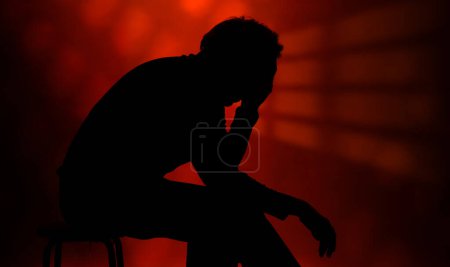 Foto de Hombre sentado, triste y pensativo, soledad y aislamiento, fondo rojo con luces que transmiten ansiedad - Imagen libre de derechos